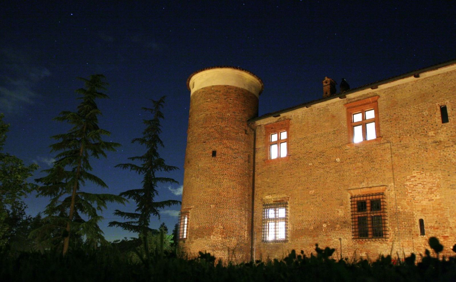 La torre del Castello Quattro Torri illuminata di notte sotto il cielo stellato, tra i cedri del parco
